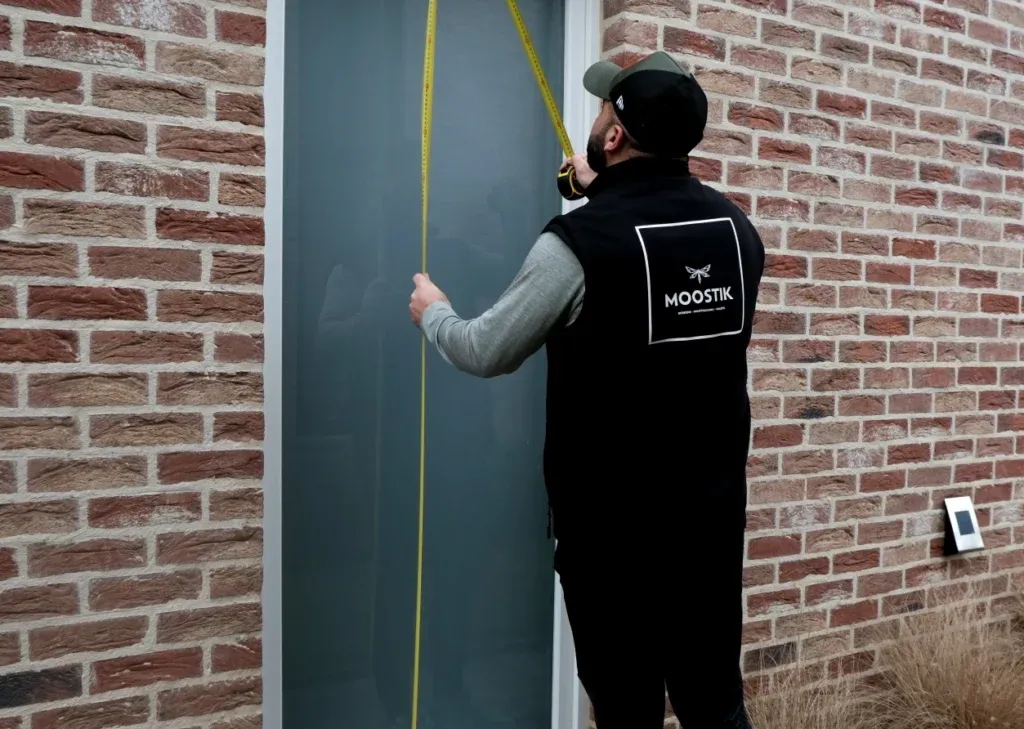 Un technicien portant une veste noire avec le logo "Moostik" mesure la hauteur d'une fenêtre avec un ruban à mesurer, devant un mur en briques rouges. Le technicien est concentré sur la prise de mesures précises pour l'installation d'une moustiquaire ou d'un volet roulant. Le mur en briques donne un aspect traditionnel à la scène, tandis que l'action met en avant le professionnalisme et l'attention aux détails nécessaires pour une installation sur mesure.