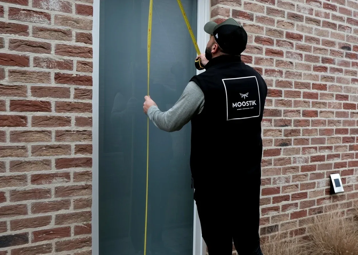 Un technicien portant une veste noire avec le logo "Moostik" mesure la hauteur d'une fenêtre avec un ruban à mesurer, devant un mur en briques rouges. Le technicien est concentré sur la prise de mesures précises pour l'installation d'une moustiquaire ou d'un volet roulant. Le mur en briques donne un aspect traditionnel à la scène, tandis que l'action met en avant le professionnalisme et l'attention aux détails nécessaires pour une installation sur mesure.